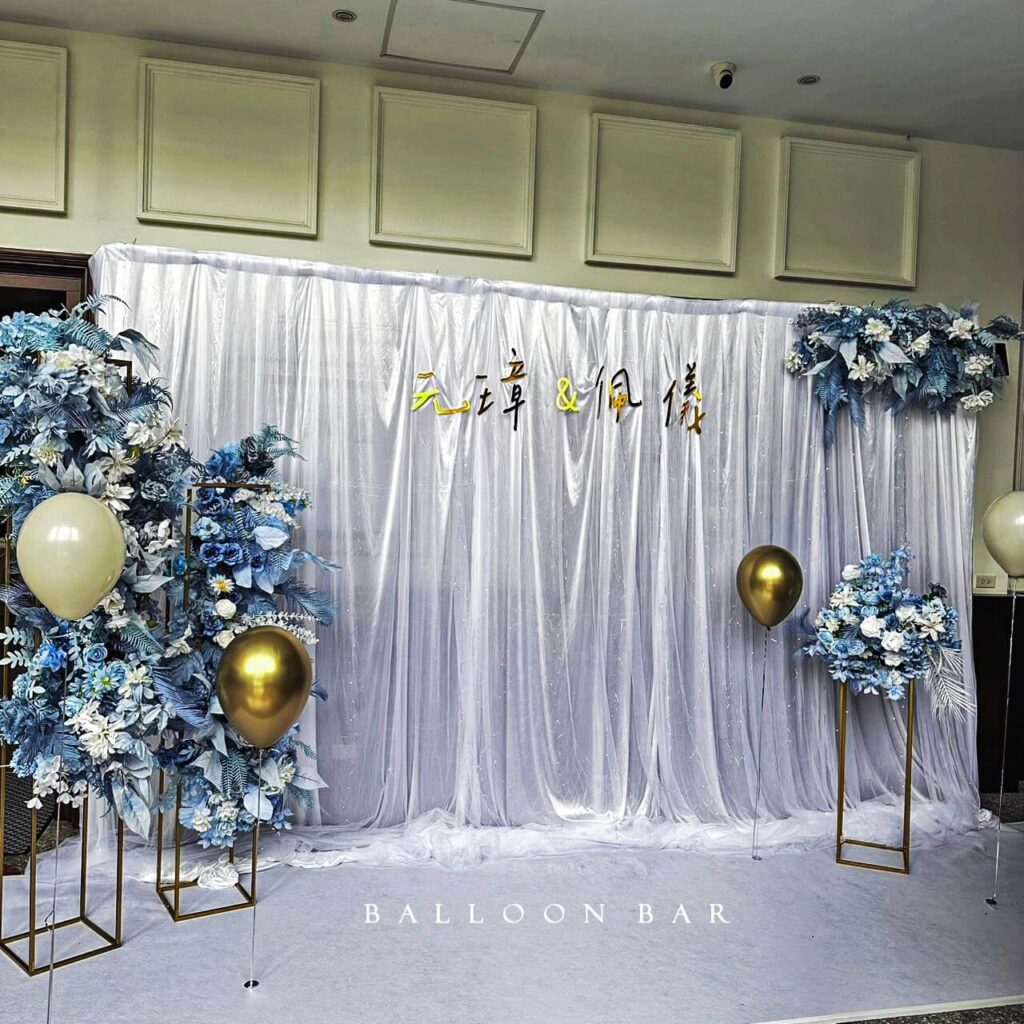 以藍色與白色為主色調，整體風格清新優雅。氣球吧團隊以半透明紗幔打造出柔和的背板層次，佈置中加入香檳金與白色氣球作為點綴，讓金屬與花材之間產生光澤對比