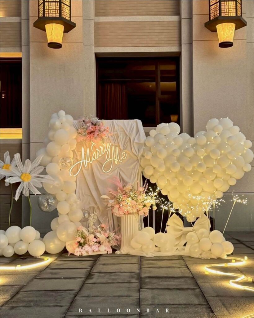 以純白與淡粉色氣球拱門搭配暖光燈串與「Marry Me」燈牌，打造出夢幻又細膩的求婚場景