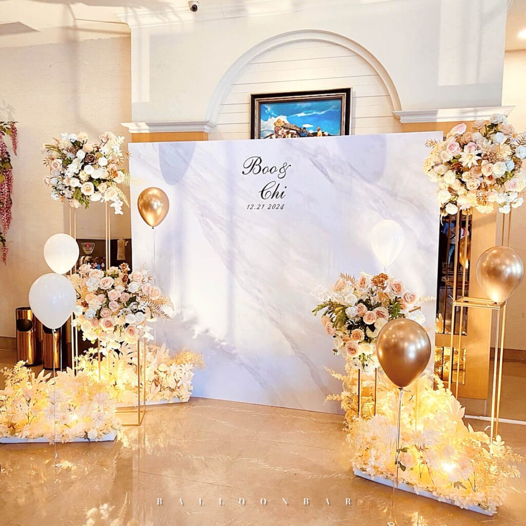 整體設計以大理石紋背板作為主視覺基底，搭配左右對稱的玫瑰花與乾燥植材花藝，並以香檳金與白色氣球點綴層次